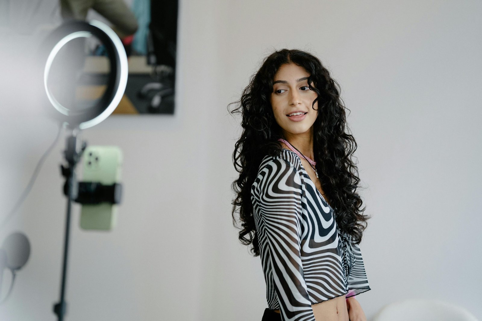 Woman with curly hair using smartphone and ring light for vlogging, wearing a patterned top.